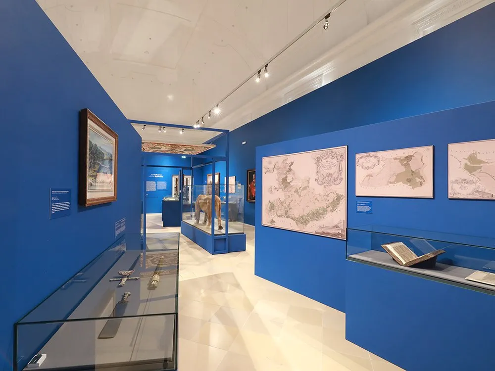 Ausstellungsraum mit blauen Wänden, gefüllt mit Glasvitrinen, die historische Gegenstände wie Schwerter und ein Buch zeigen, sowie Wandkarten und ein Gemälde