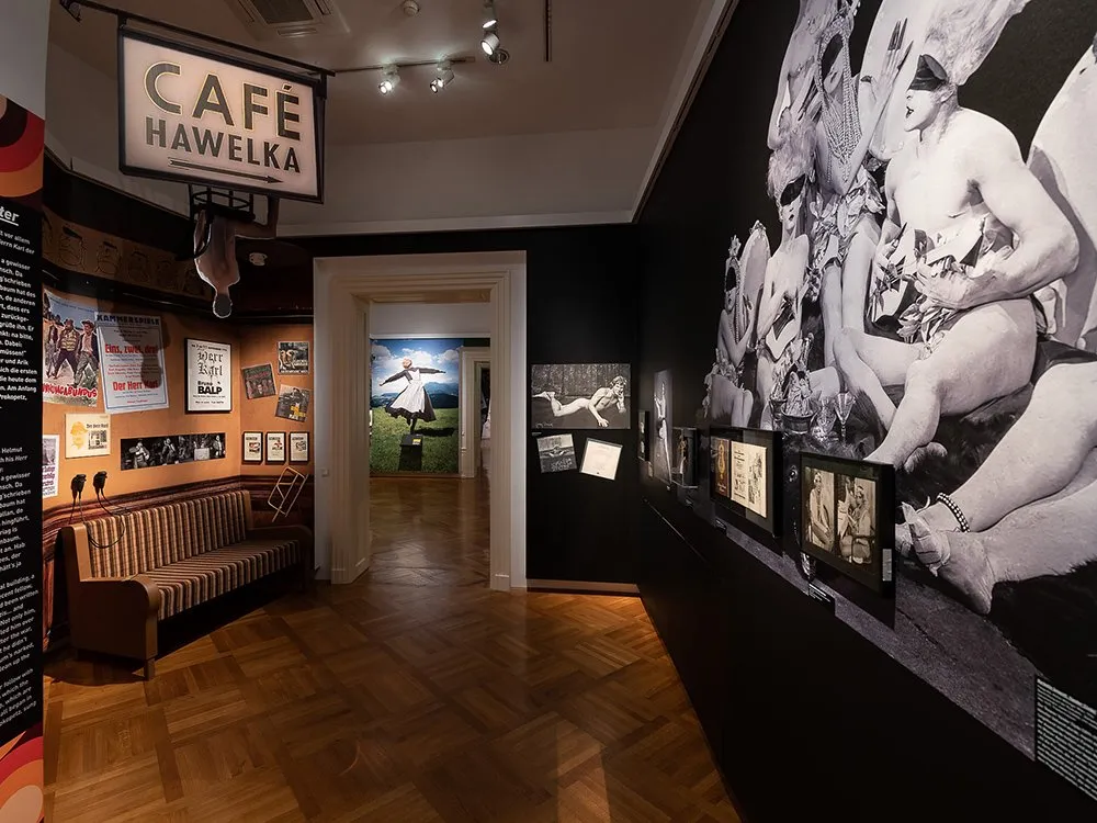Ausstellungsraum: "Café Hawelka" Schild über einem braunen Sofa. An der Wand hängen Poster und Fotos.