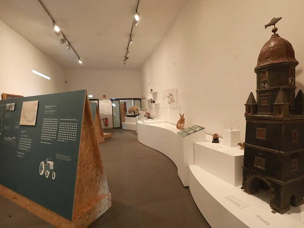 Ausstellungsraum: weiße wellenförmige Ausstellungsfläche mit Tierfiguren und Turmmodell mit Fischwetterfahne, grünes Schaukelbrett mit Karte und Traktor