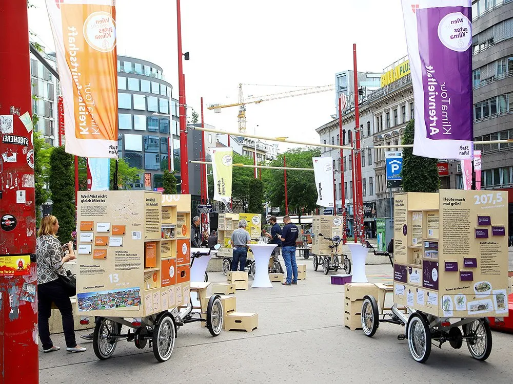 Auf einer Straße stehen E-Lastenfahrräder mit Holzkisten und Infos zu Klimathemen. Im Hintergrund sieht man Gebäude und Fahnen“. Menschen betrachten die Fahrräder.
