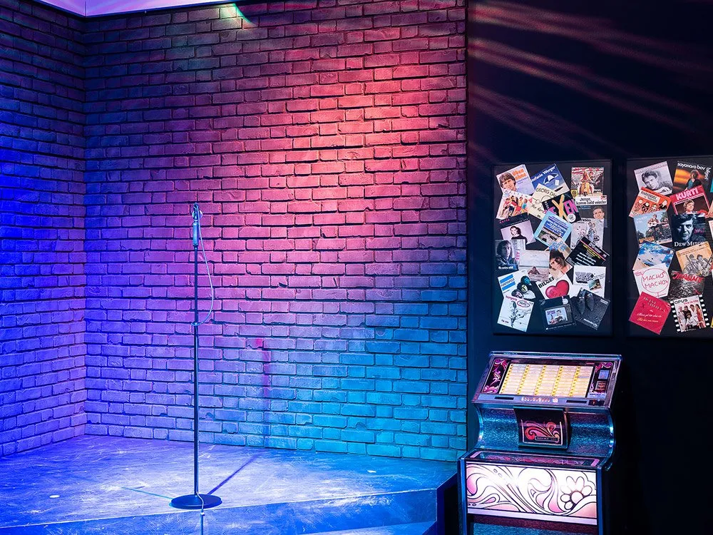 Bühne mit einer Ziegelwand, beleuchtet in Pink- und Blautönen, und einem Mikrofonständer in der Mitte. Rechts steht ein bunter Jukebox mit Verzierungen.
