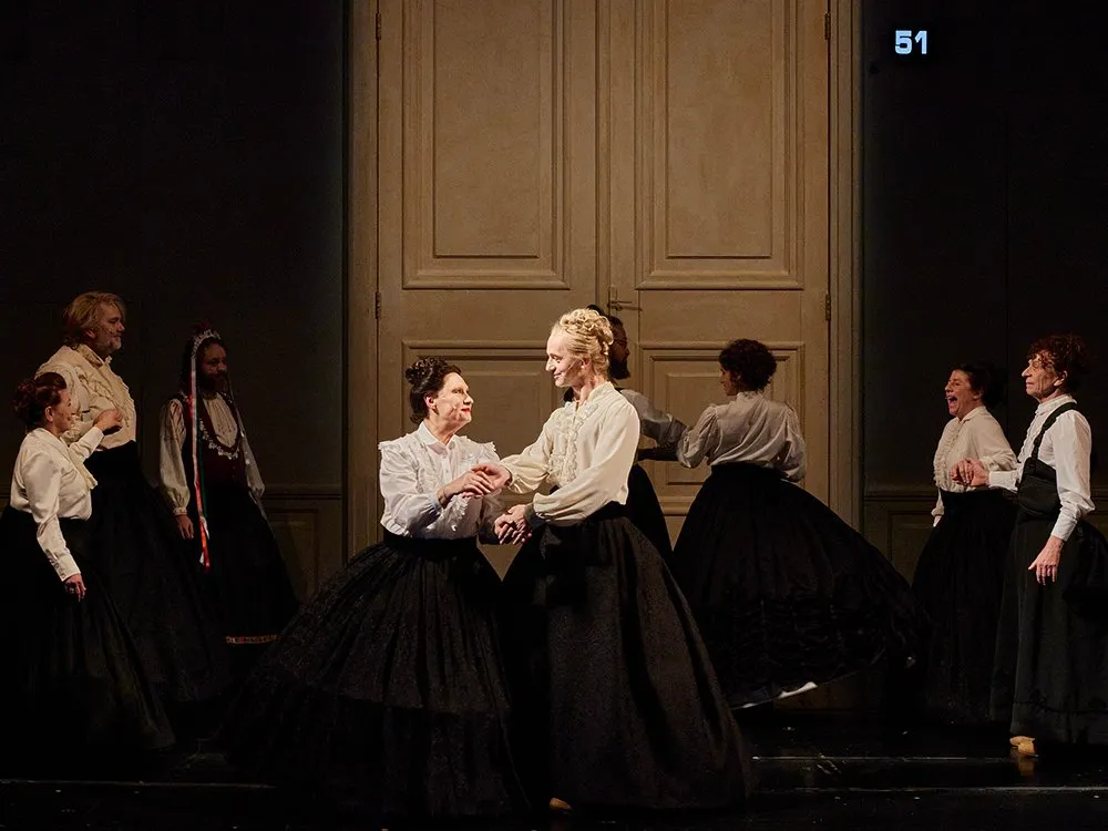 Szene: Darsteller in historischen schwarzen und weißen Kostümen tanzen vor einer großen Holztür