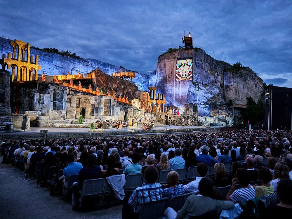 Bühnenbild mit Darstellern in farbigen Kostümen, die sich vor einem goldenen Tempel und steinernen Häusern bewegen, umgeben von einer lichterbesetzten Landschaft.