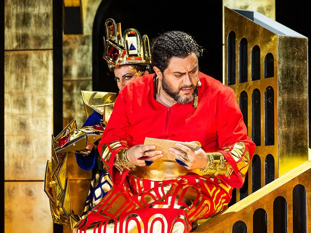 Szene: Mann in rot-goldenem Gewand mit Buch auf Treppe, Person im blau-goldenen Kostüm mit Krone im Hintergrund