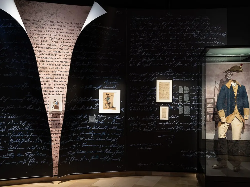 Schwarze Wand voller handschriftlicher Texte. In der Mitte ein offenes Buch mit Schriften und einer kleinen Figur. Rechts eine Vitrine mit einer alten Uniform und Dokumenten.