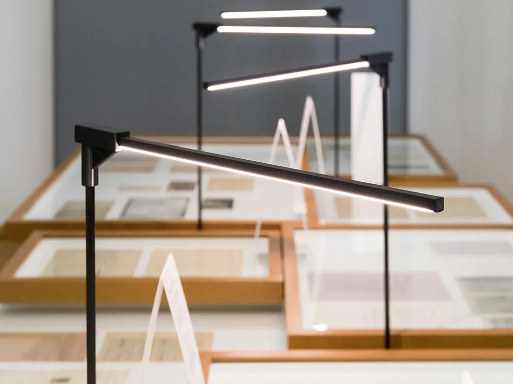 Detailansicht: Moderne, schlanke Lampen mit langen Leuchtstreifen über Tischen mit Vitrinen und Dokumenten.