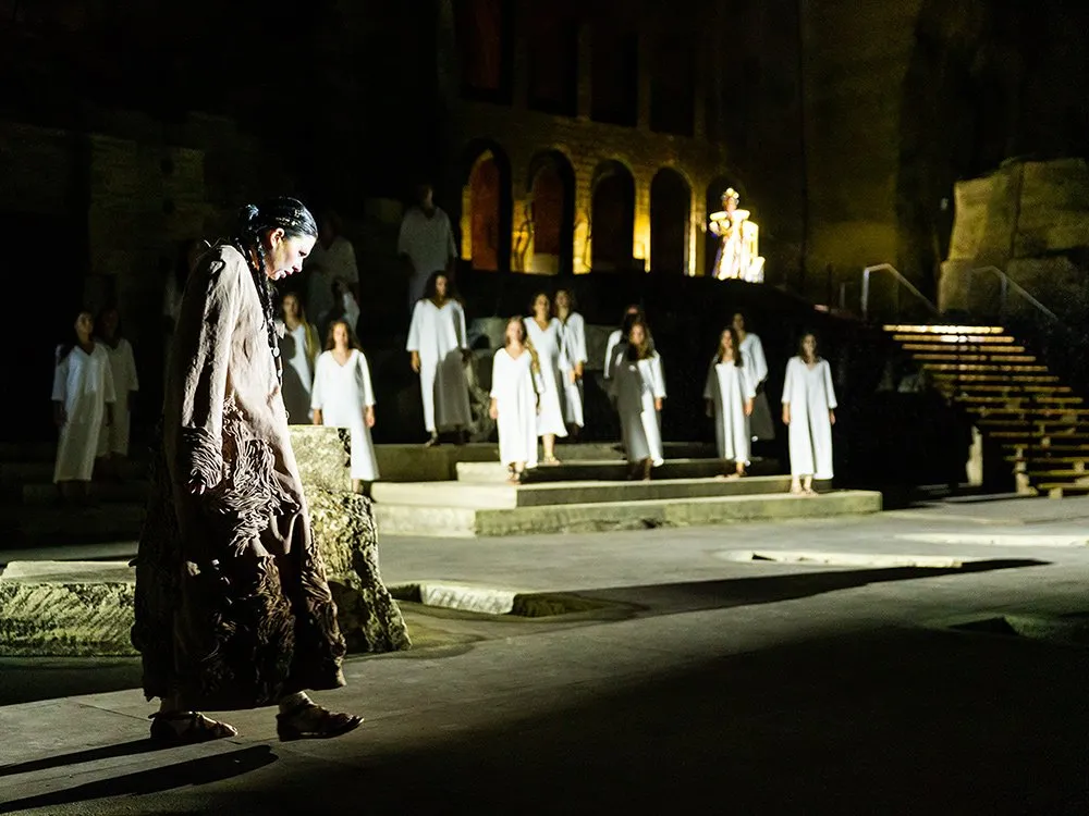 Szene: Person in braunem Gewand vorne, Gruppe in weißen Roben auf Treppe im Hintergrund
