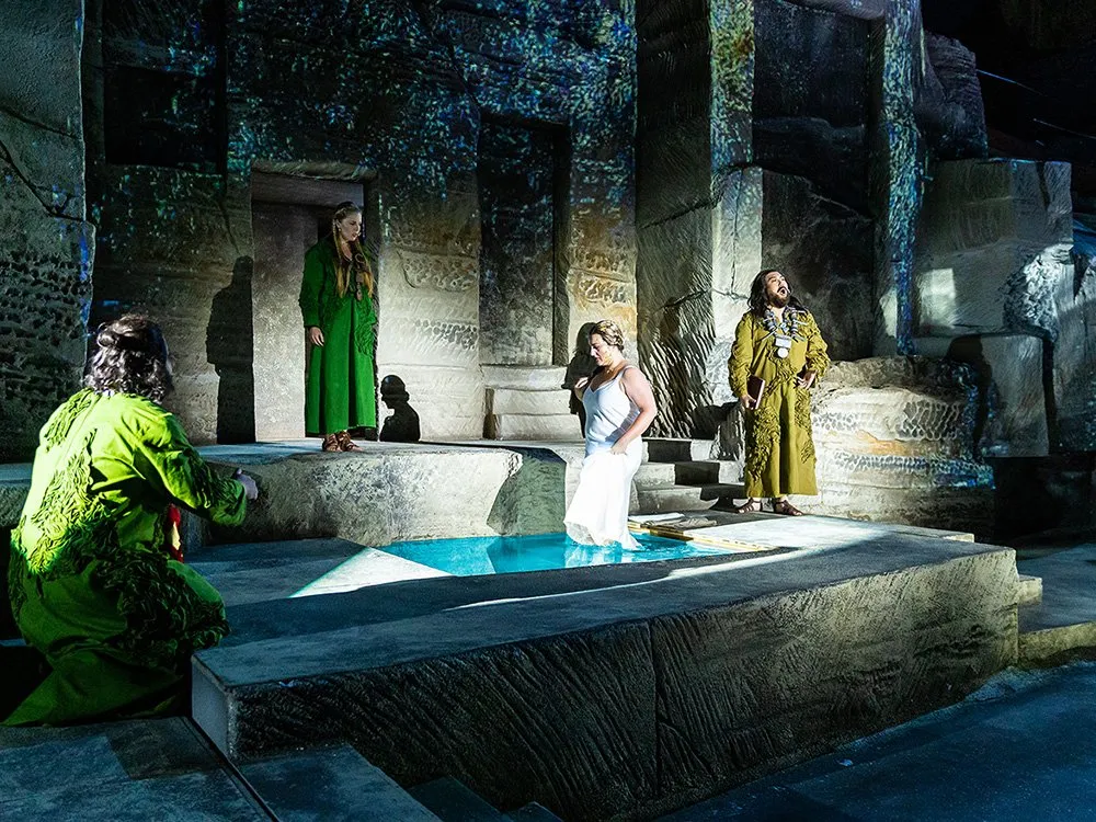 Szene: Darstellerin steht in einem Wasserbecken, umgeben von zwei anderen, eine kniend und eine stehend, vor steinerner Kulisse mit Bögen.