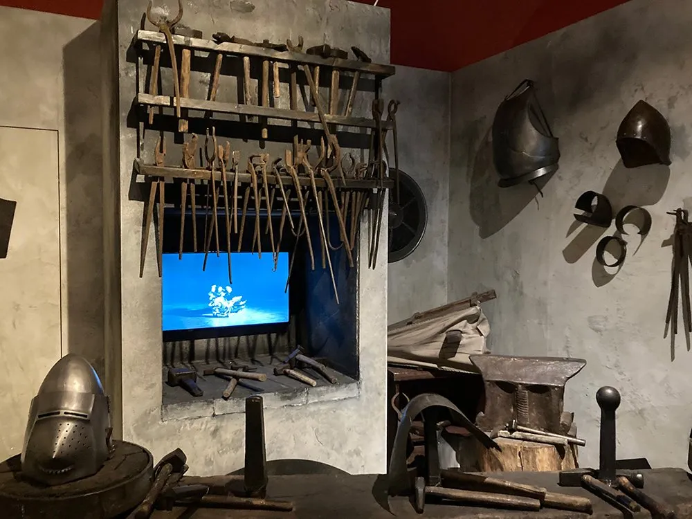 Mittelalterlicher Raum mit Regal voller alter Werkzeuge, einem Amboss, einem Helm und einem Bildschirm, an einer steinernen Wand.