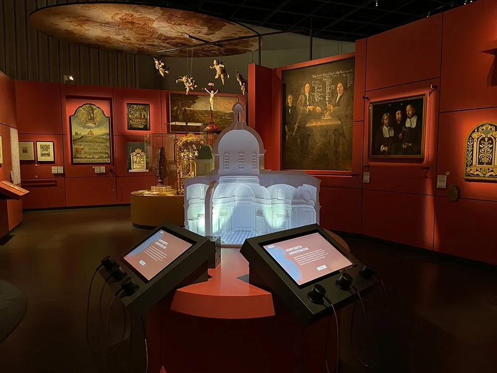 Ausstellungsraum mit roten Wänden, Modell einer Kirchenhälfte und interaktiven Touchscreens, umgeben von historischen Gemälden und hängenden Engelfiguren.