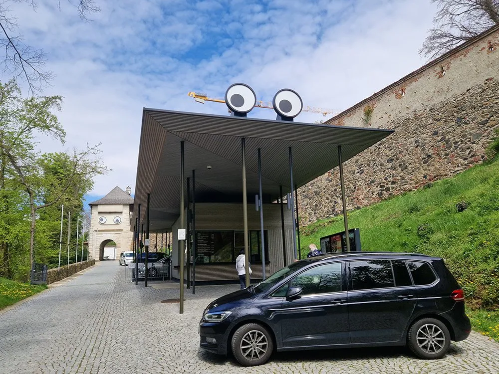 Aufgang zur Schallaburg mit überdimensionalen Augen auf dem Dach und einem weiteren Augenpaar am Turm, neben geparkten Autos und einer steinernen Mauer.