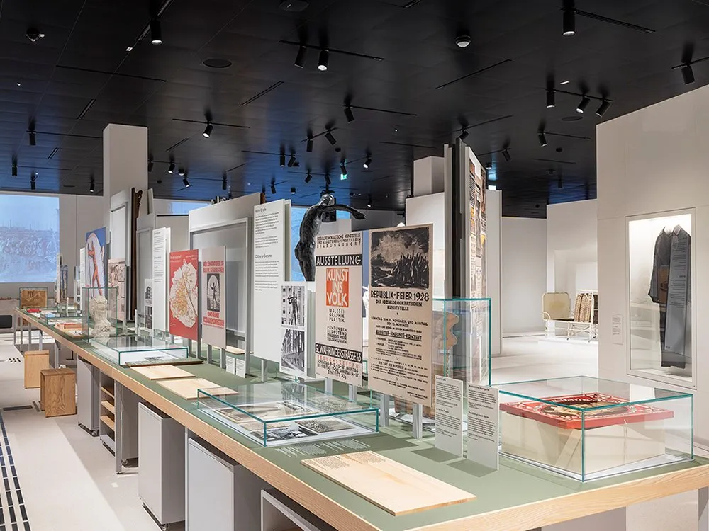 Ausstellungsraum mit einer Reihe von Vitrinen und Tischen, die historische Dokumente, Plakate und Kunstwerke zeigen