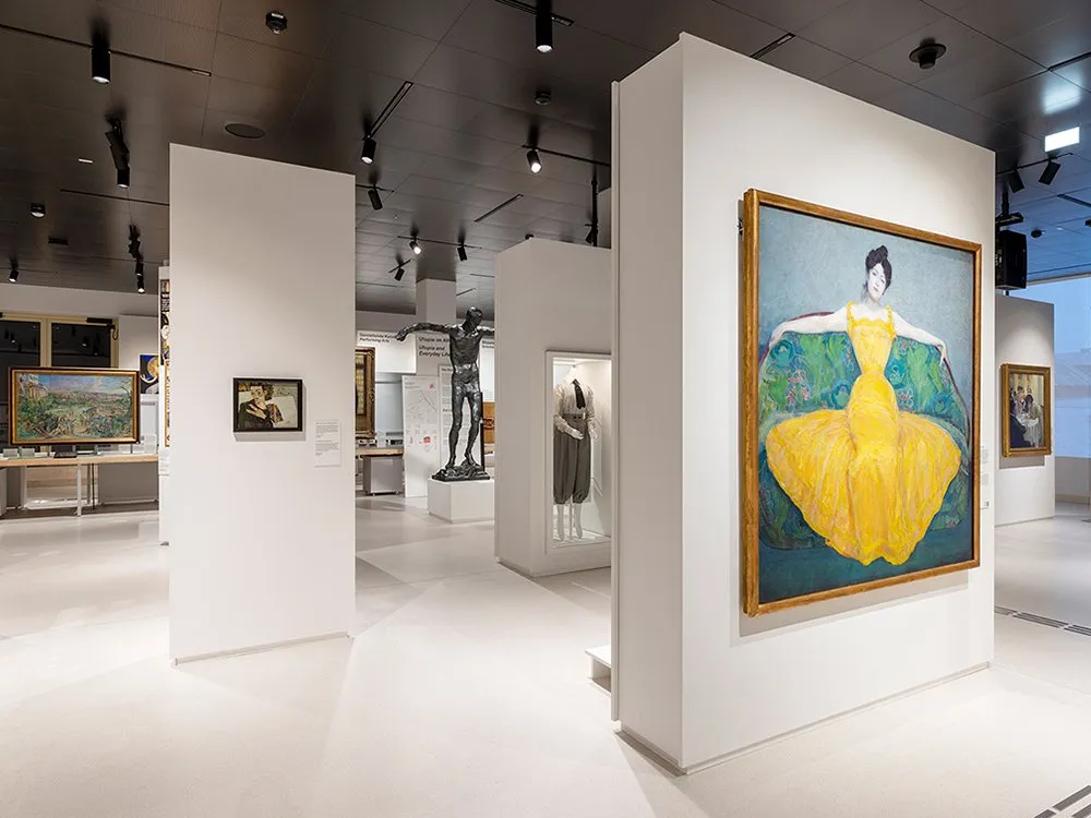 Ausstellungsraum mit einem großen Gemälde einer Frau in gelbem Kleid, Vitrinen mit Statuen, Kleidung und anderen Exponaten