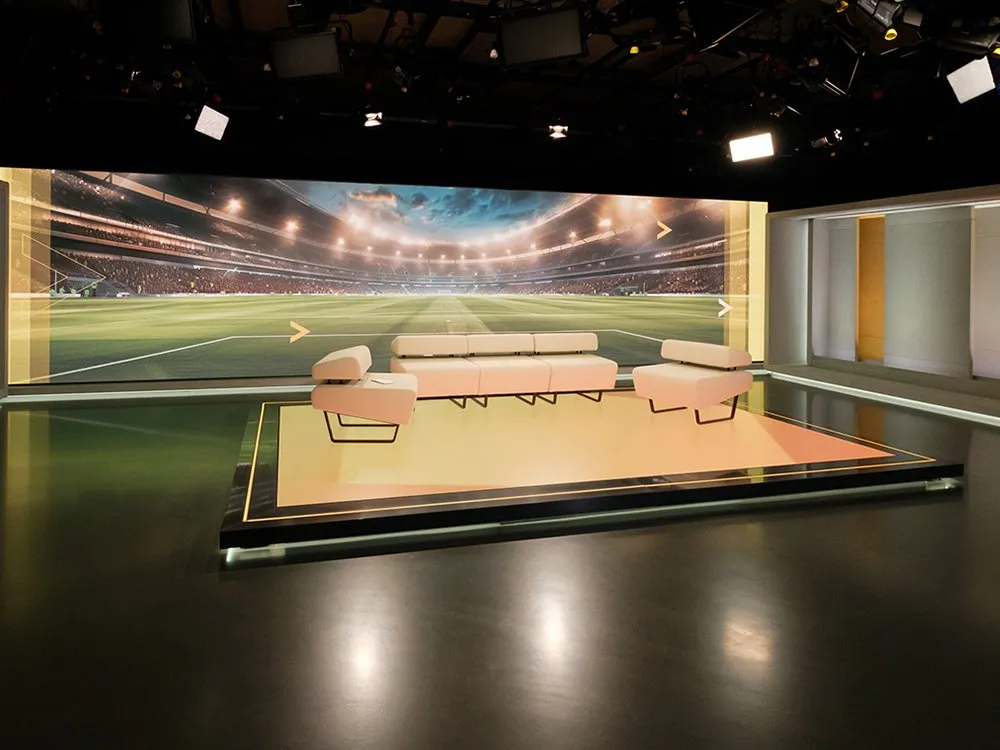 Modernes Sportstudio mit großer LED-Rückwand, die ein Stadion zeigt, umgeben von Sitzmöbeln auf einem beleuchteten Podest.