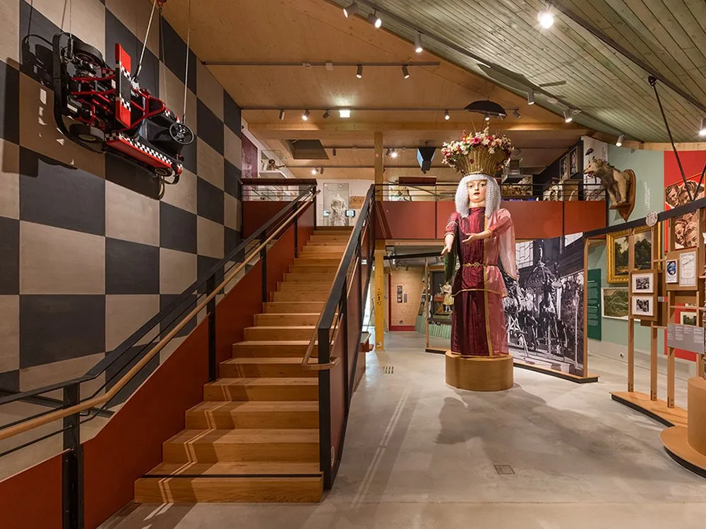 Innenraum des Pratermuseums mit einer zentralen Fortuna-Figur auf einem Drehteller, einem GoCard-Modell, das von der Decke entlang der Wand hängt, und einer schachbrettgemusterten Wand. Eine Treppe führt zu einer Galerie mit weiteren Ausstellungsstücken.