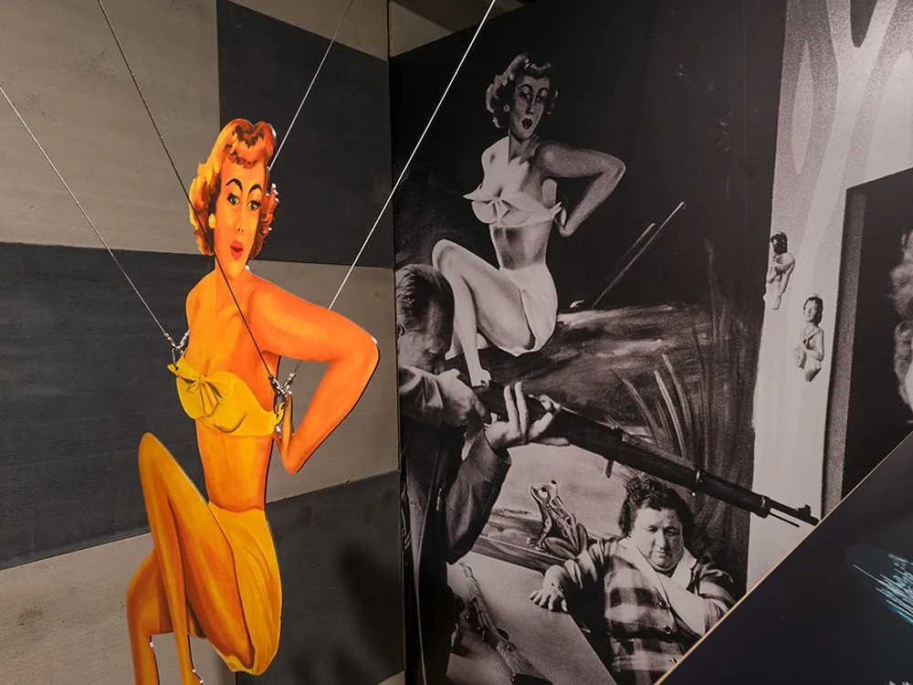 Ausstellungsbereich:  gelbe Pin-Up-Figur, die von der Decke hängt, und einem Schwarz-Weiß-Foto von einer Szene mit einer Person und einem Gewehr, vor einer schachbrettgemusterten Wand.