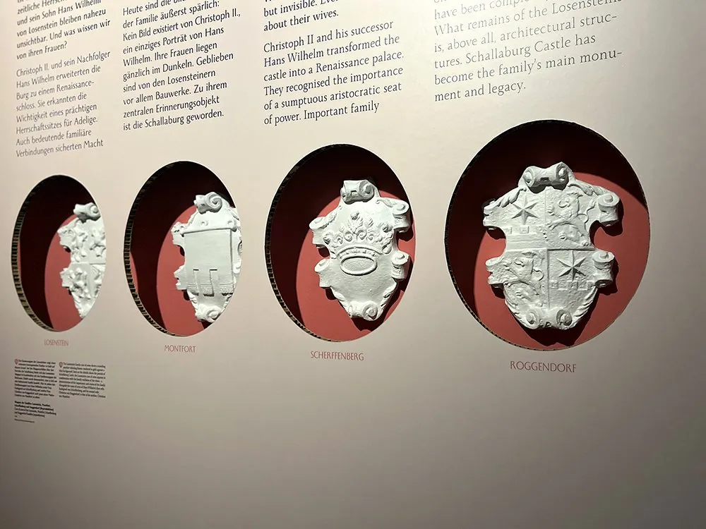 Vier weiße Wappenmodelle (Losenstein, Montfort, Scherffenberg, Roggendorf) hängen an einer roten Wand. Daneben sind Texte über die Familien und die Schallaburg.