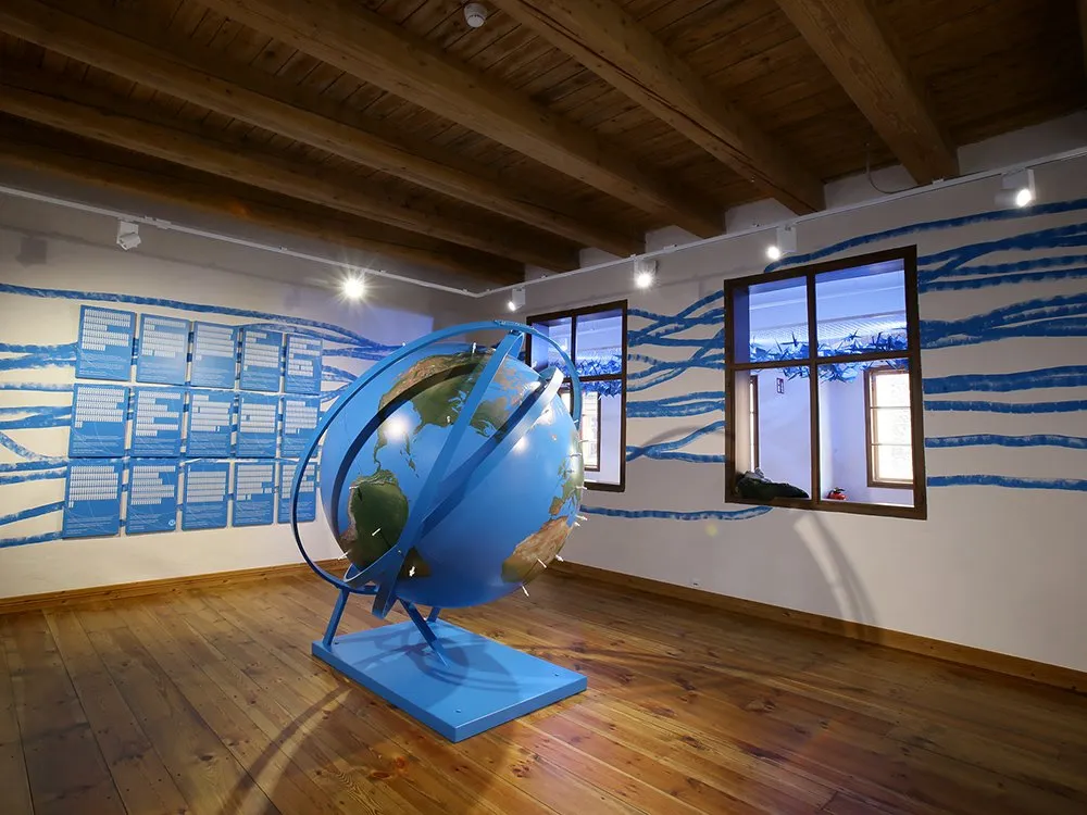 Ausstellungsraum mit einer großen blauen Erdkugel im Zentrum, umgeben von thematischen Wänden mit Listen und blauen Wellenmustern.