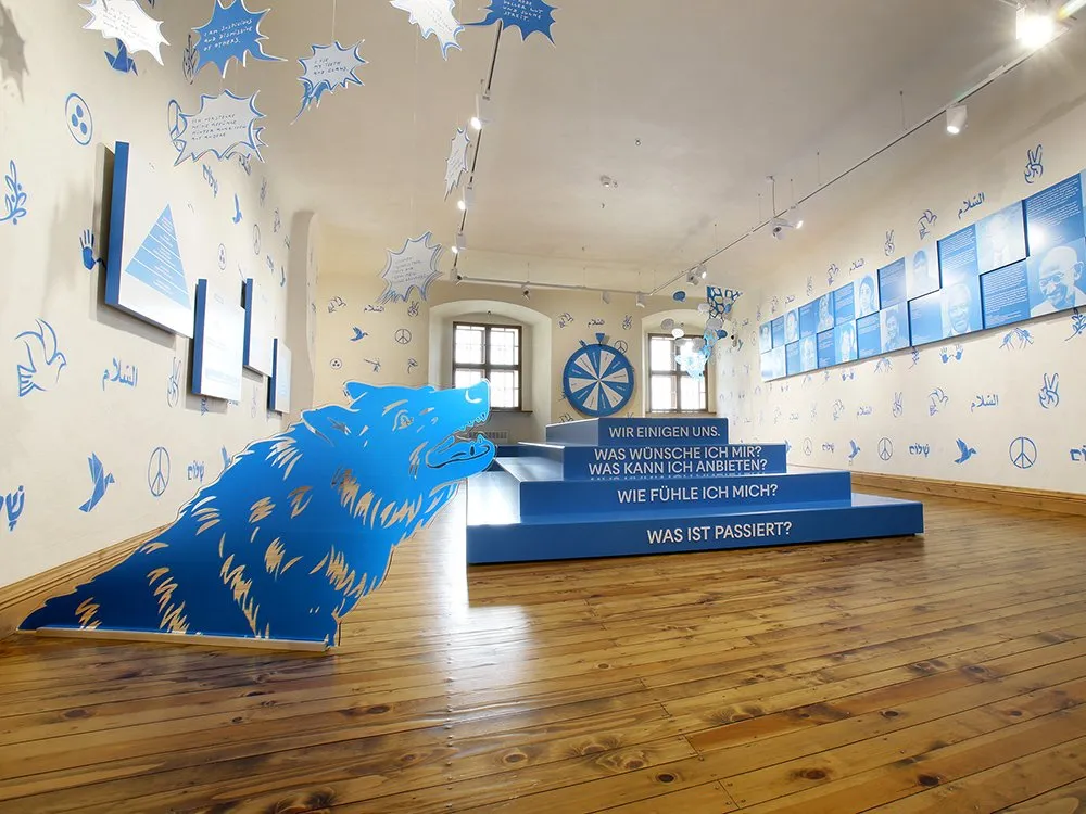 Ausstellungsraum mit blauen Podesten und Texten, einem großen Wolfsymbol am Boden, hängenden Schildern und dekorativen Wänden mit Friedenssymbole