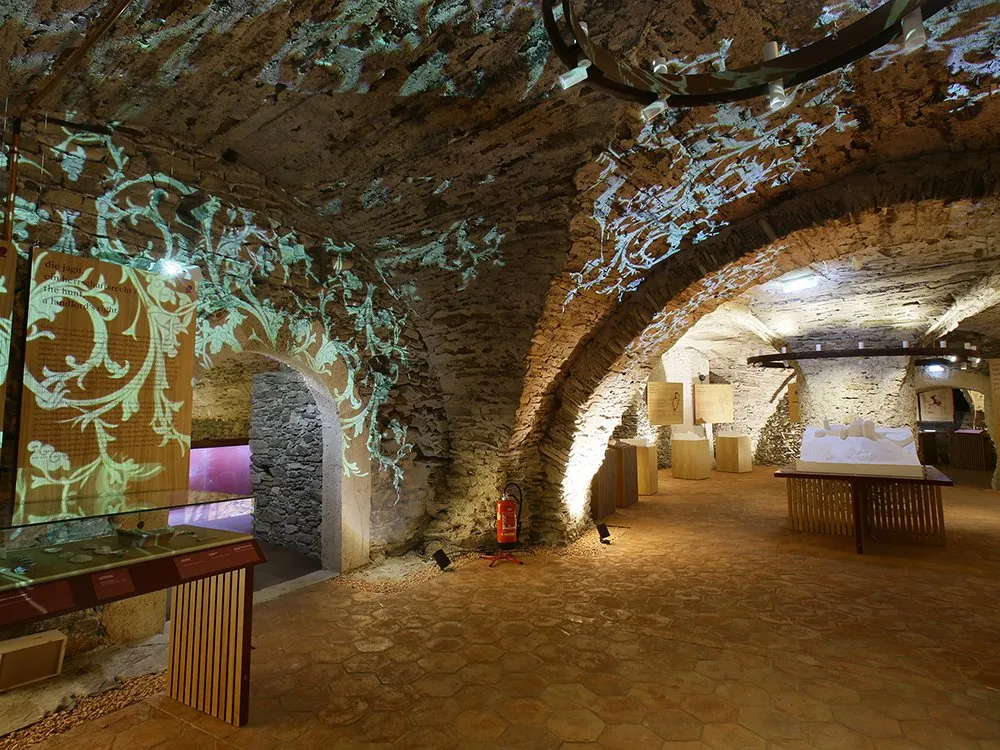 Ausstellungsraum mit gewölbten Steinwänden, beleuchtet mit projizierten Ornamenten, und Ausstellungsstücken auf Vitrinen und Tischen