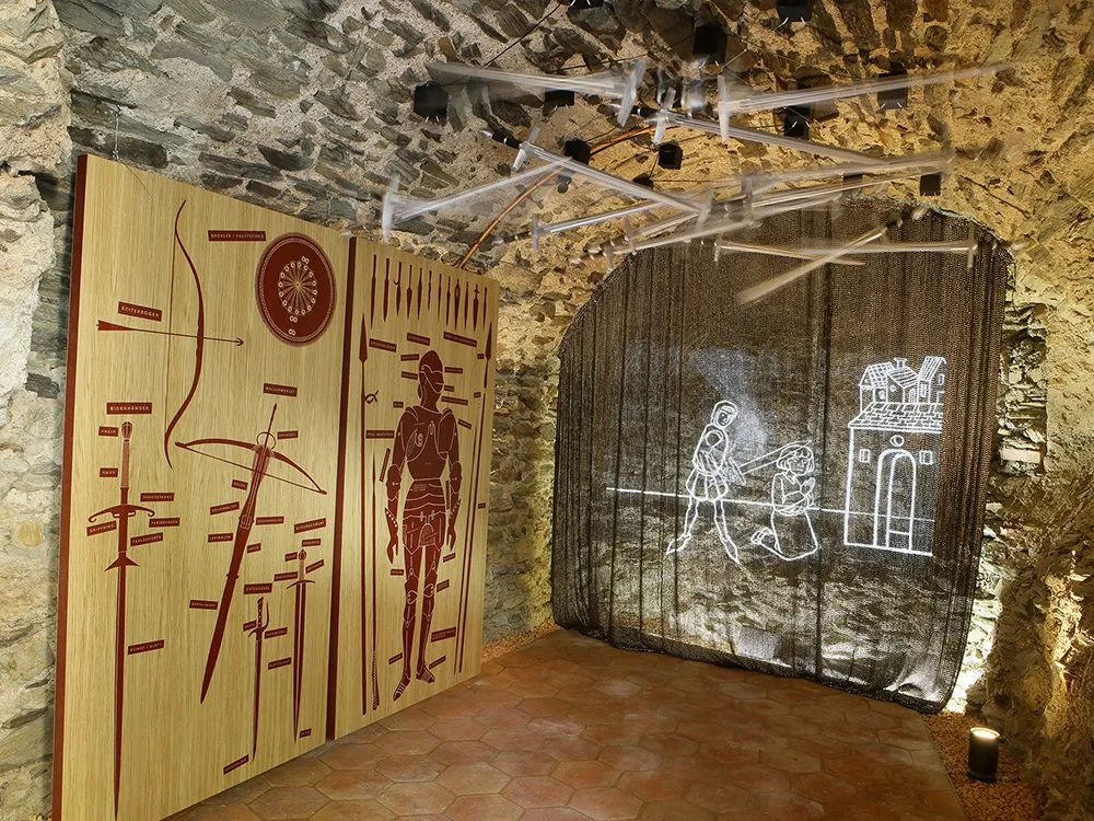 Ausstellungsraum mit steinernen Gewölben, beleuchteten Panels mit Waffendarstellungen und einer projizierten Szene mit kämpfenden Figuren.