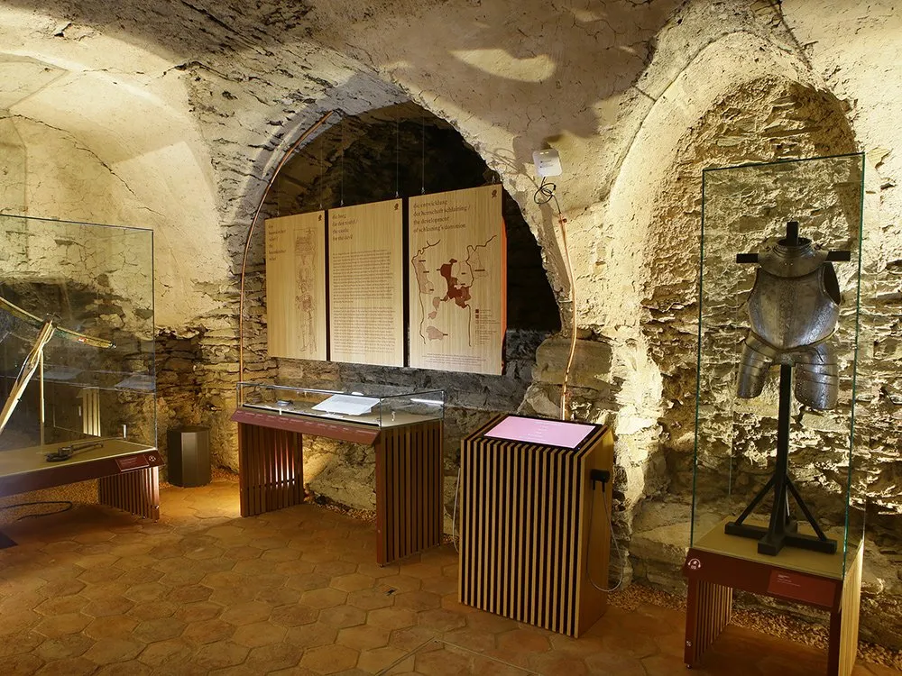 Ausstellungsraum mit steinernen Gewölben, beleuchteten Karten an der Wand, Vitrinen mit Rüstungen und einem interaktiven Display.