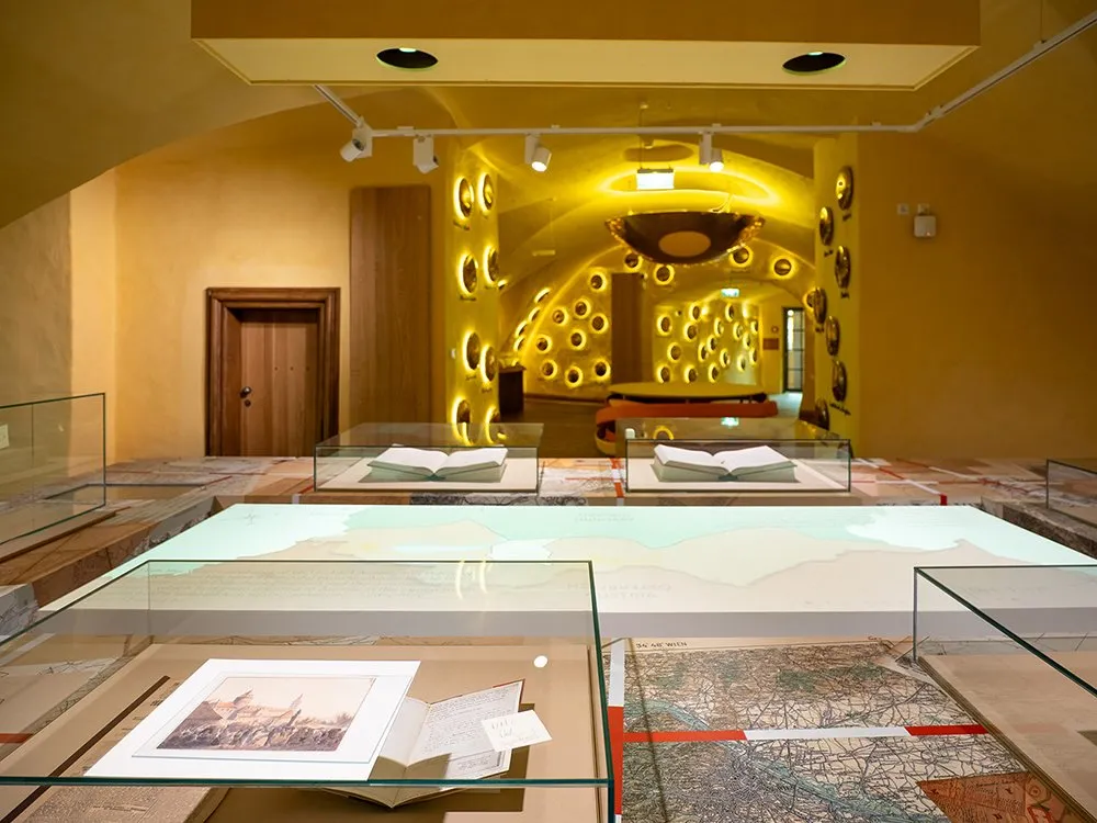 Ausstellungsraum mit beleuchteten Wänden mit runden Elementen und Vitrinen mit Karten und historischen Dokumenten. 5,2 s