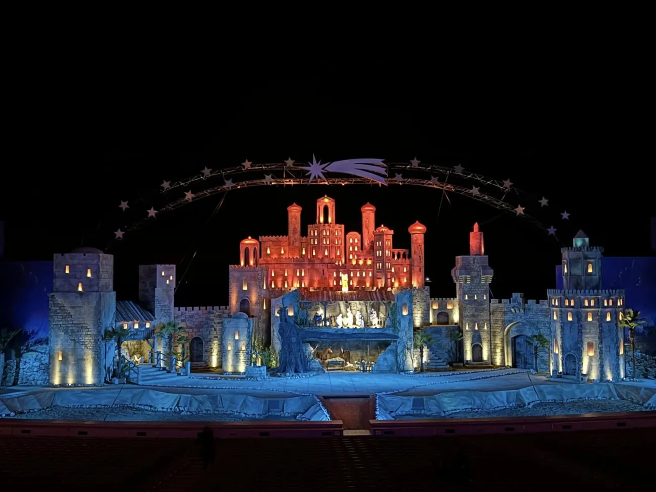 Beleuchtete Theaterbühne, mit steinernen Türmen, Gebäuden in rotem Licht beleuchtet und einem Bogen voller Sterne und strahlenden Sternschnuppe.