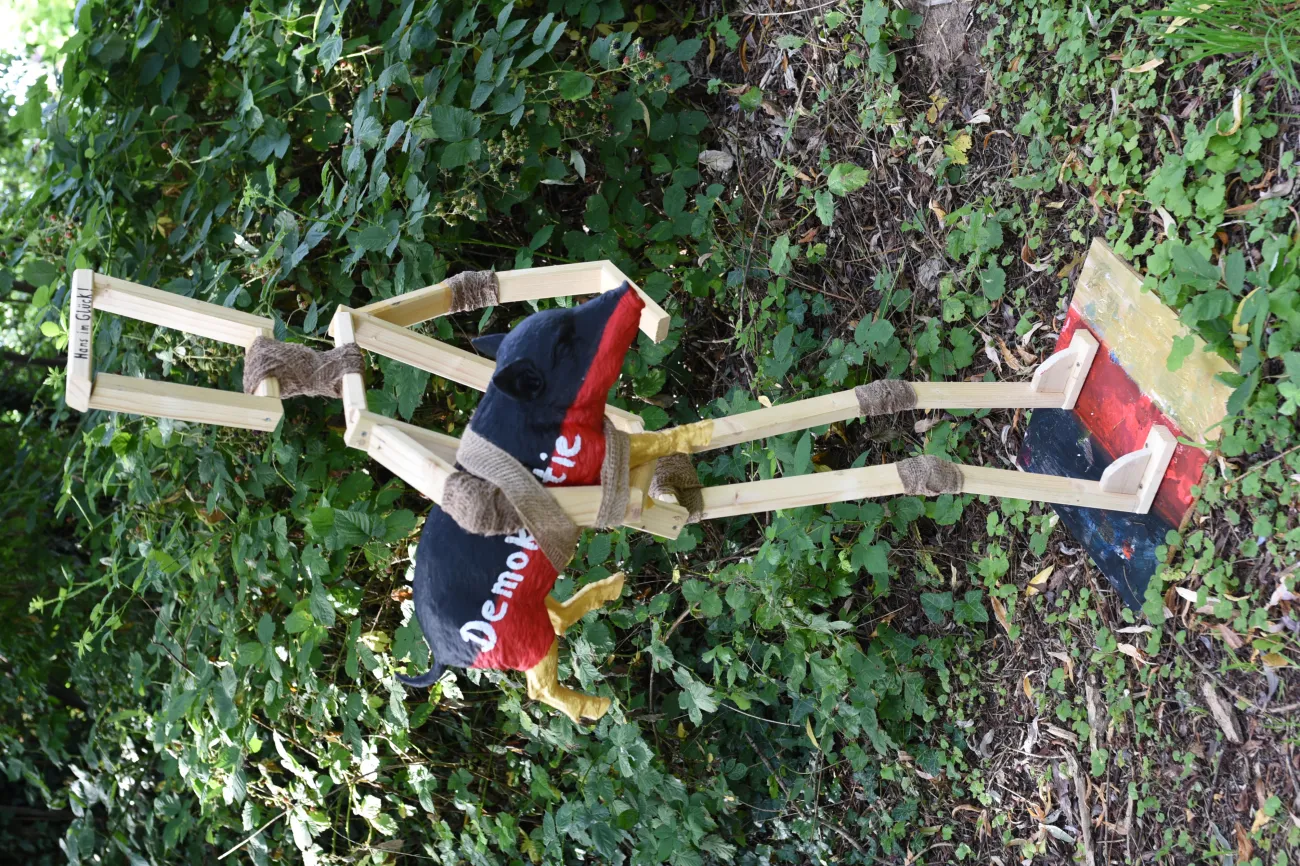 Holzmännchen hält Schwein mit Aufschrift "Demokratie" bemalt wie Deutschlandfahne