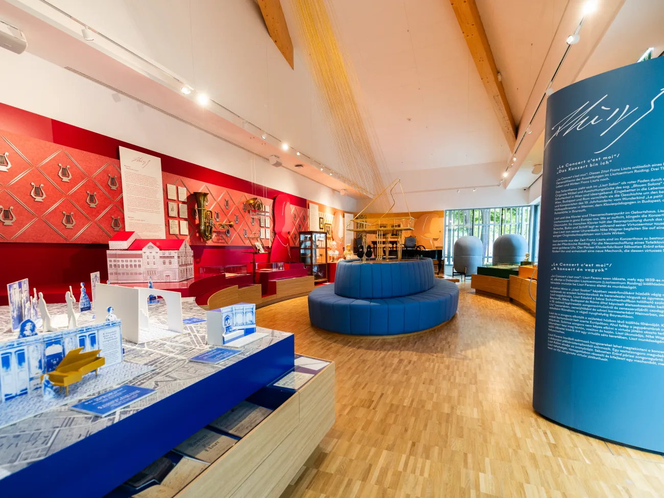 Moderner Ausstellungsraum. Es gibt ein Modell einer historischen Stadt mit Figuren, Wände mit Musikinstrument-Dekorationen und Informationspaneelen. In der Mitte steht eine runde blaue Sitzfläche mit Kopfhörern, auf denen man Kompositionen von Liszt hören kann, sowie ein Klavier auf der Plattform.