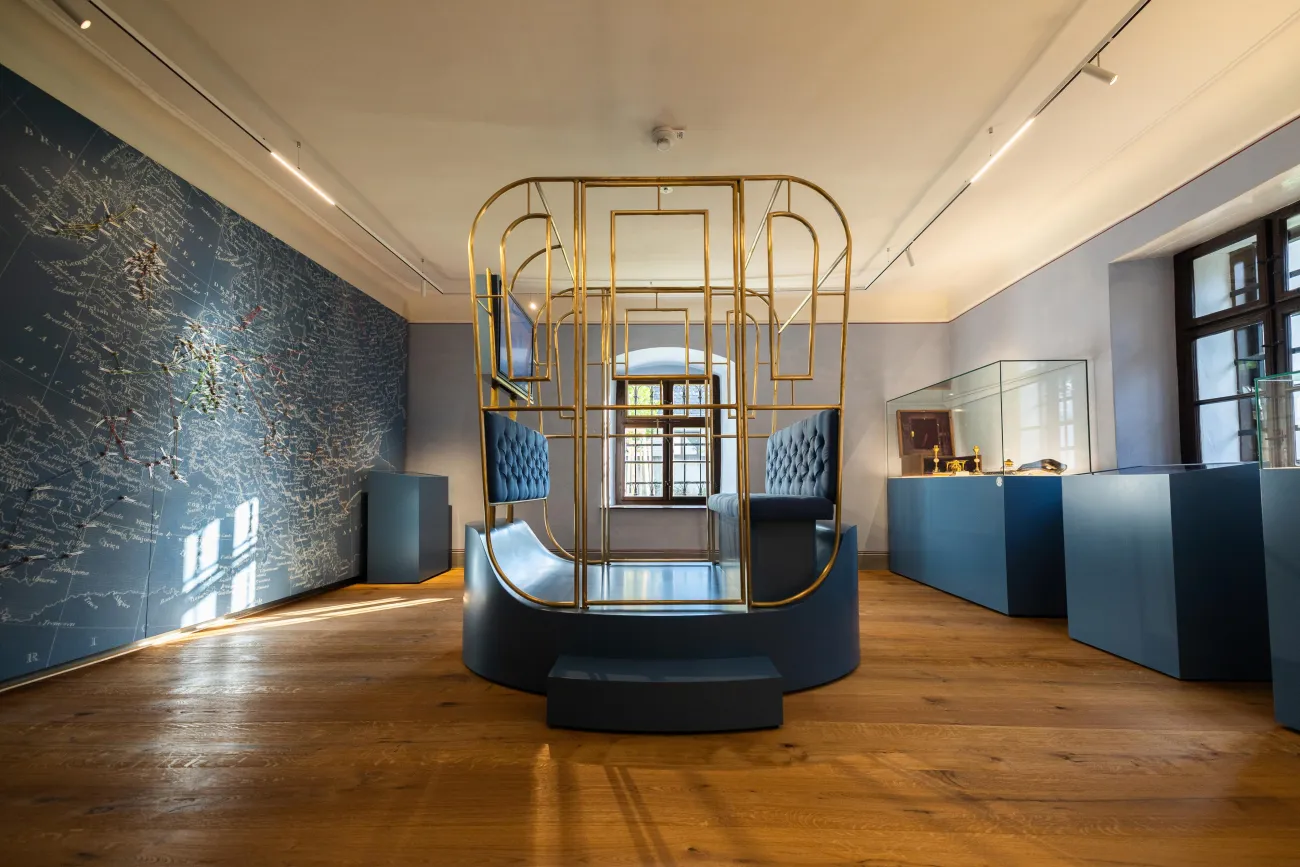Ausstellungsraum: Ein Modell einer Kutsche in der Mitte mit gepolsterter Sitzfläche und einem Monitor. An den Wänden eine Europa-Karte und Vitrinen mit Exponaten.