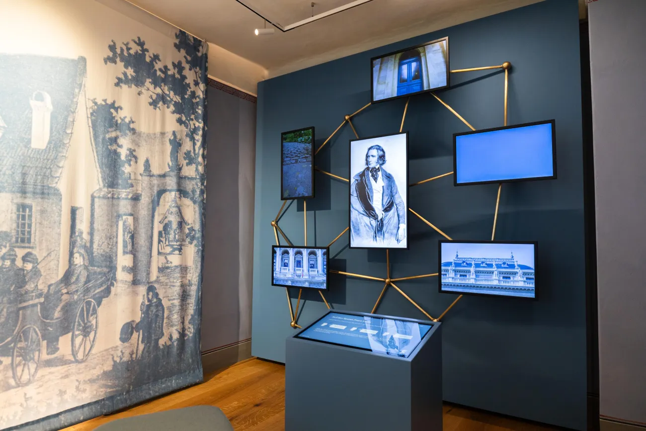 Ausstellungsraum: Installation mit einem Touchscreen-Pult in der Mitte, umgeben von mehreren Bildschirmen, angebracht an einem goldenen, geometrischen Gestell. Im Hintergrund ein Wandteppich mit historischen Szenen.