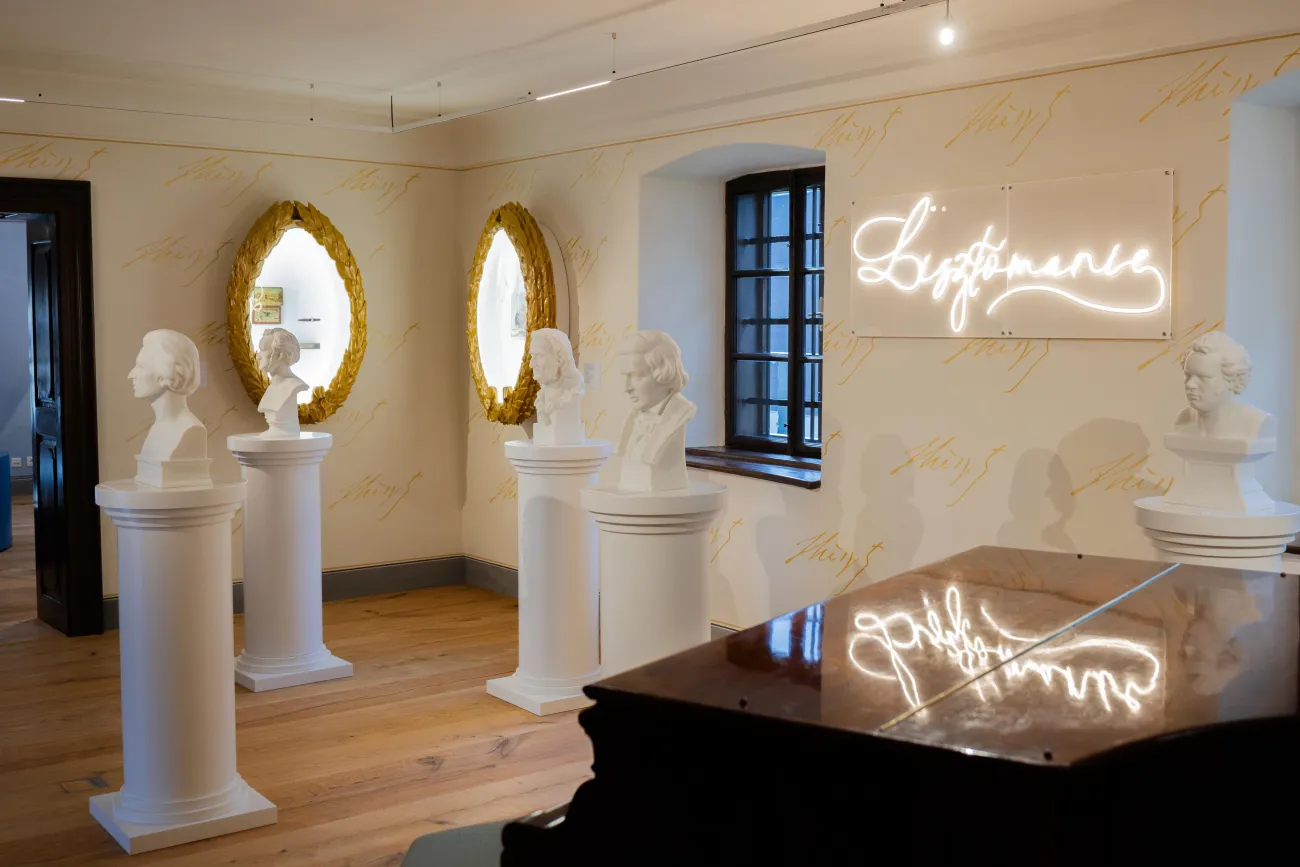 Ausstellungsraum: Mehrere weiße Büsten auf Säulen. An der Wand beleuchtete Rahmen mit Exponaten und ein leuchtender Schriftzug "Lisztomanie". Im Vordergrund ein Klavier, auf der Oberfläche spiegelt sich die Leuchtschrift.