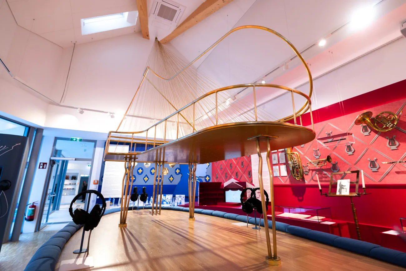 Detailansicht: großes, goldenes Klavier-Modell. Von der Decke sind goldene Saiten gespannt, die zum Klavier führen, darunter befinden sich Kopfhörer zum Anhören von Musik.