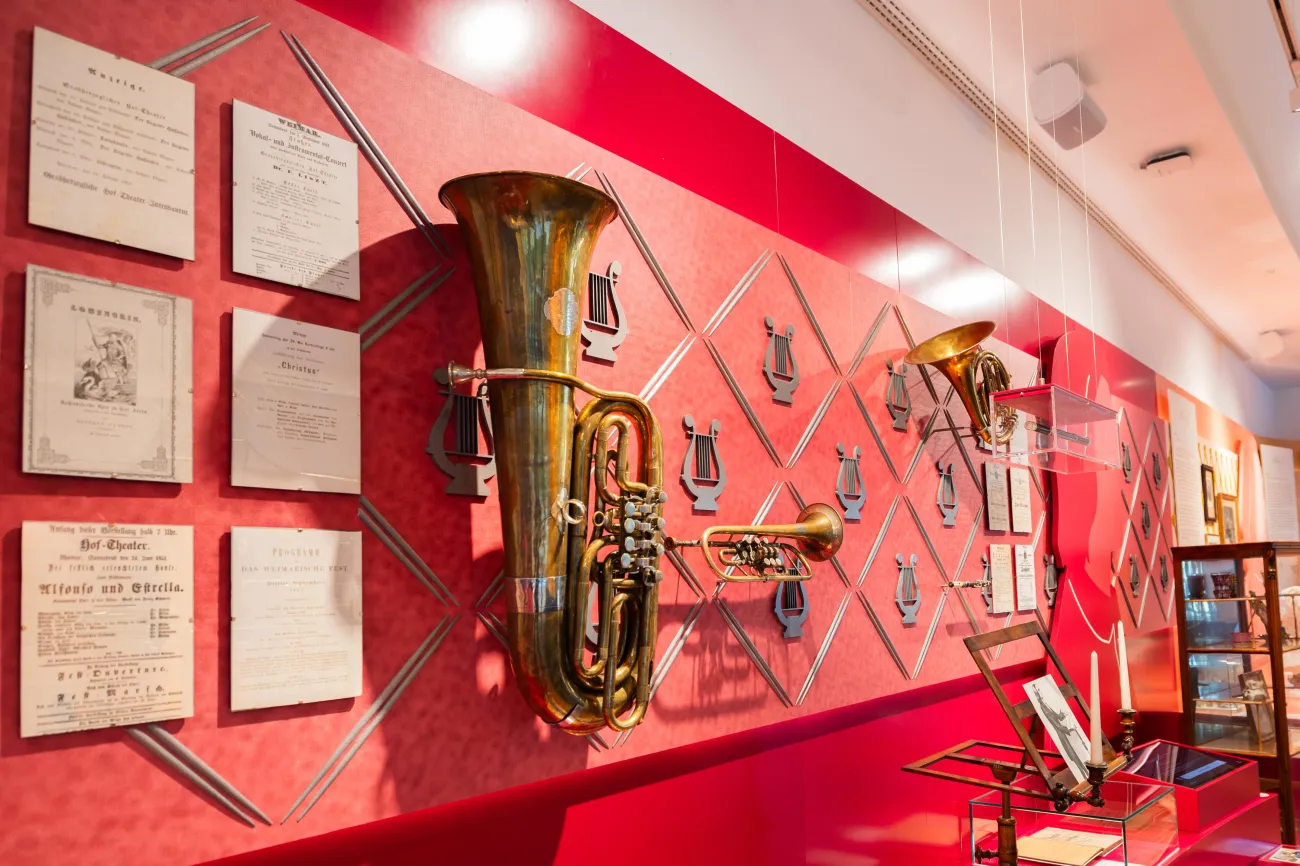 Detailansicht: Wand geschmückt mit Blasinstrumenten. Dazwischen sind gerahmte Dokumente und Notenblätter angebracht, die historische Informationen über Franz Liszt bieten.