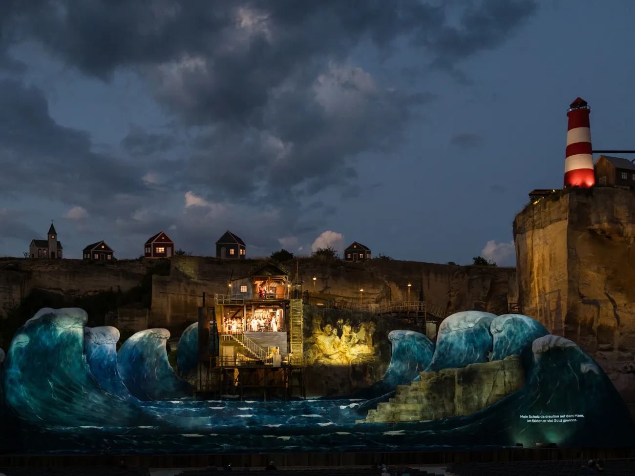 Nächtliche Freiluftbühne, hohe künstliche Wellen, mittig ein beleuchtetes Holzhaus auf Stelzen, darin Schauspieler. Dahinter steile Felsen mit kleinen Häusern, rechts oben ein rot-weißer Leuchtturm. Projektionen auf Wellen und Fels.