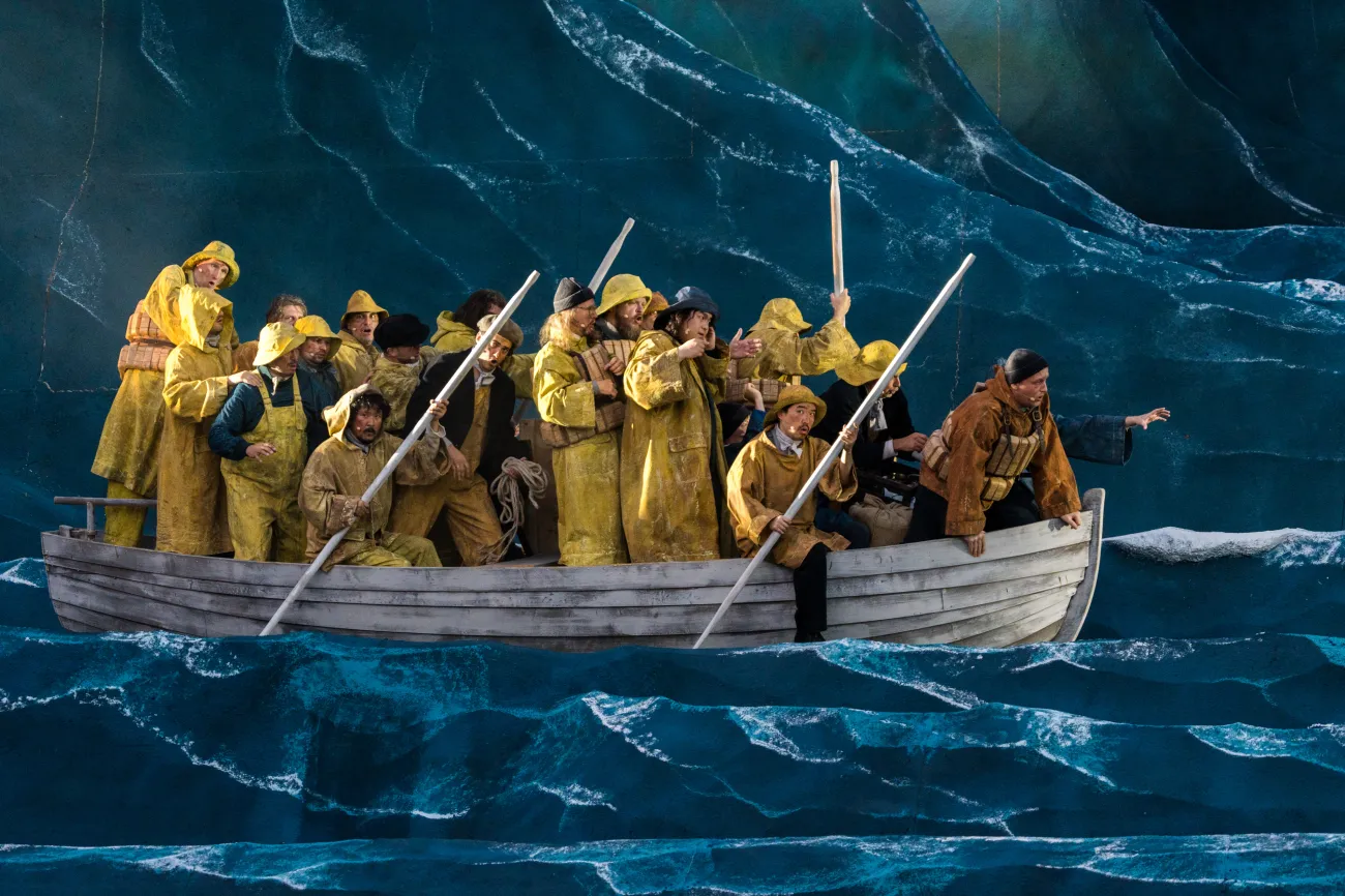 Szene: kleines Holzboot, vollgepackt mit Darstellern in gelben Regenmänteln inmitten von großen Wellen