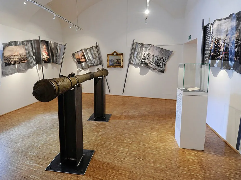 Ausstellungsraum: Antike Kanone zentral im Raum, Fahnen aus Metall mit Bildern und Text an Wänden