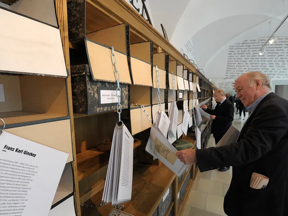 Holzregal mit Fächern, beschriftet mit Namen. Darin hängen an Ketten Dokumente und Papiere, die von Personen in Anzügen betrachtet werden