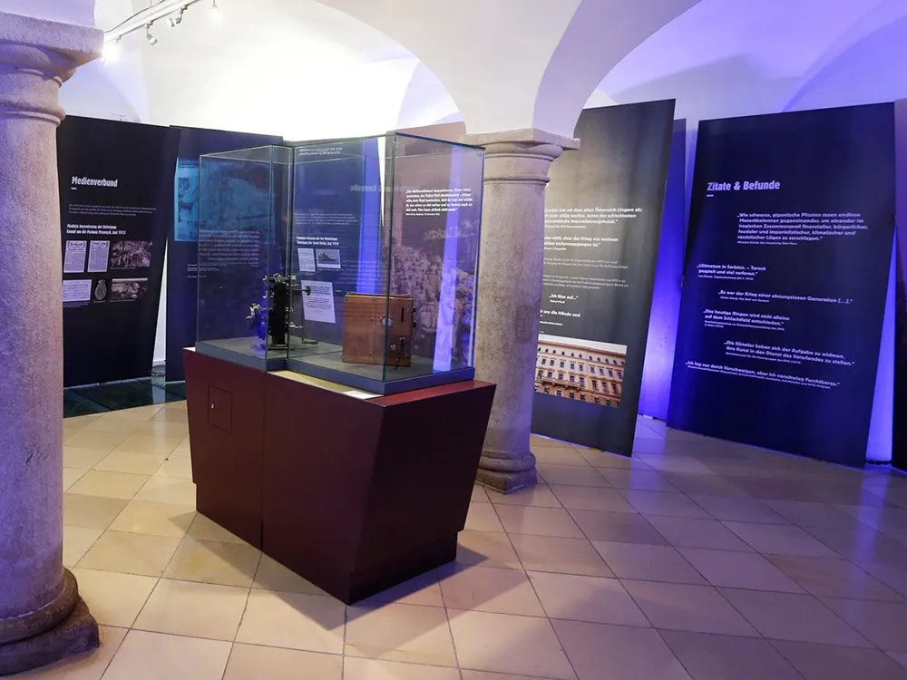 Gewölbter Raum mit Säulen, beleuchtet in Blau. Im Zentrum eine Vitrine mit Ausstellungsstücken, umgeben von Infotafeln