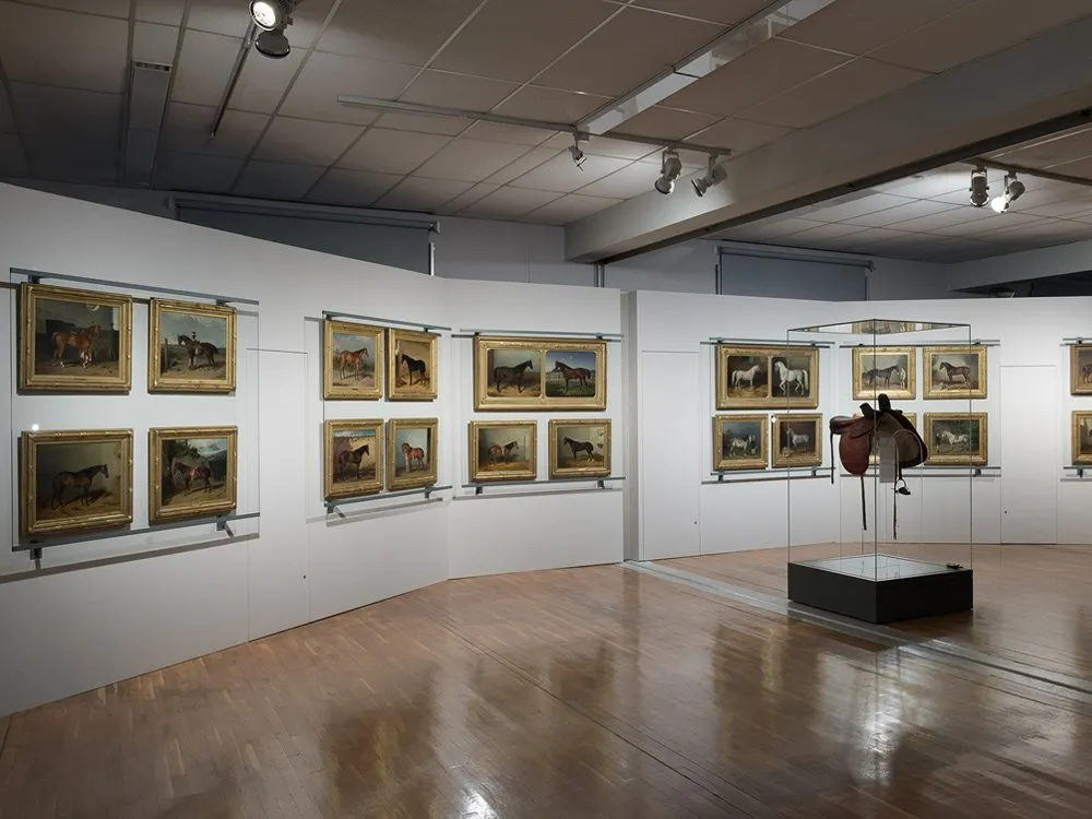 Ausstellungsraum: Wände mit Gemälden von Pferden und eine Vitrine mit einem Sattel