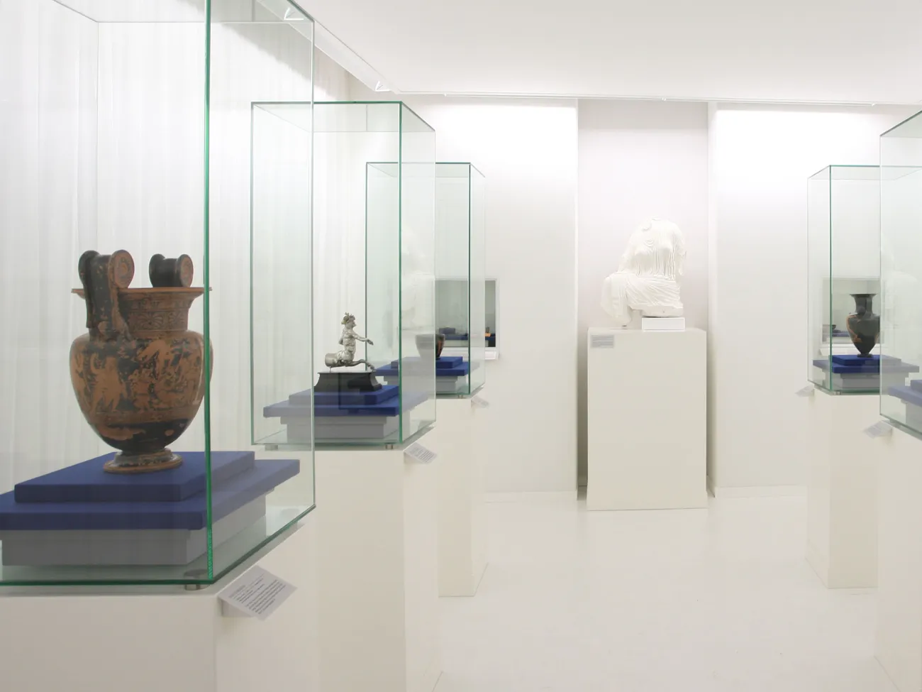 Ausstellungsraum: weißer Raum, Glasvitrinen mit Antiken Amphoren, zentral weiße unvollständige Skulptur einer Frau