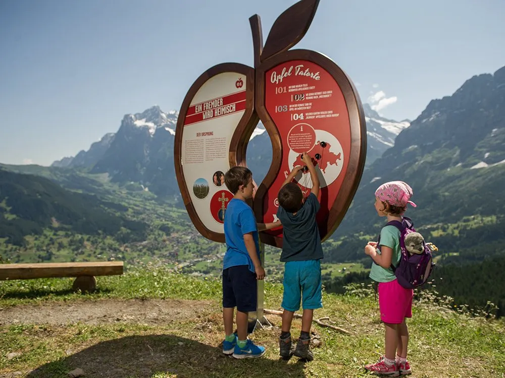 Drei Kinder stehen vor einem großen, apfelförmigen Informationsschild mit Bergen im Hintergrund