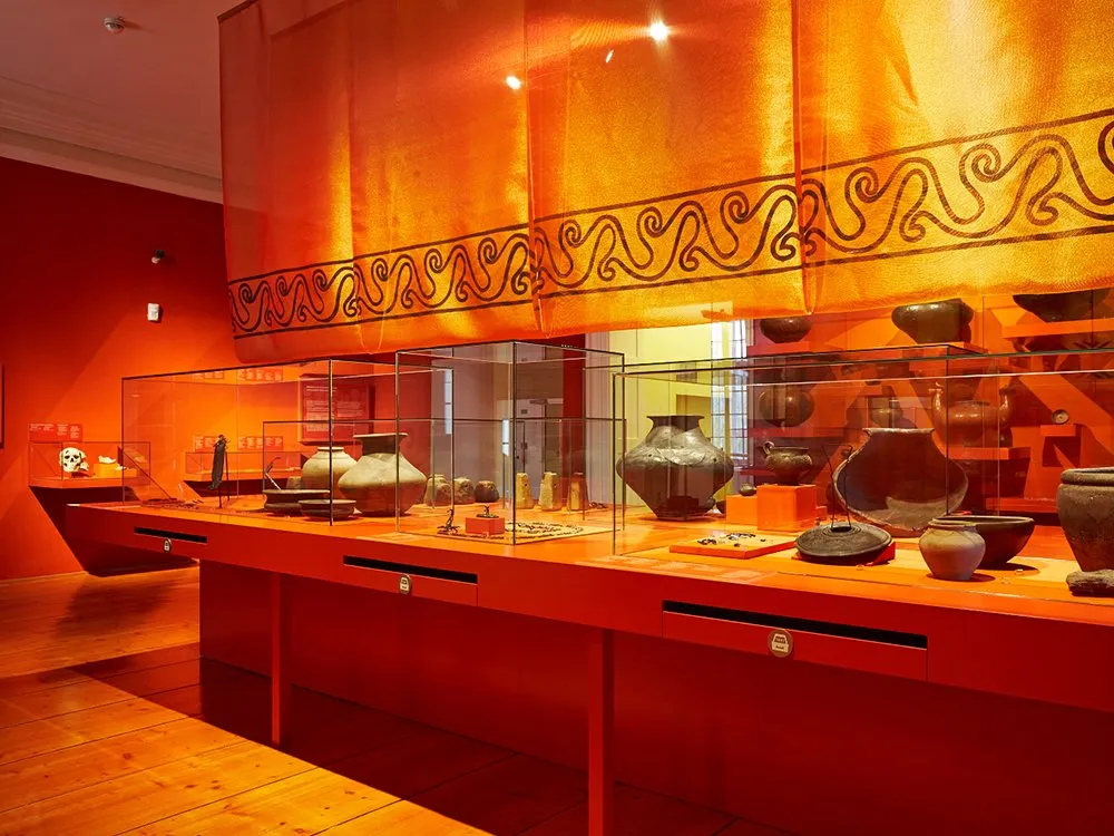 Ausstellungsraum mit Vitrinen, die Artefakte und ein Modell eines Tempels zeigen, sowie einer roten Säule im Hintergrund