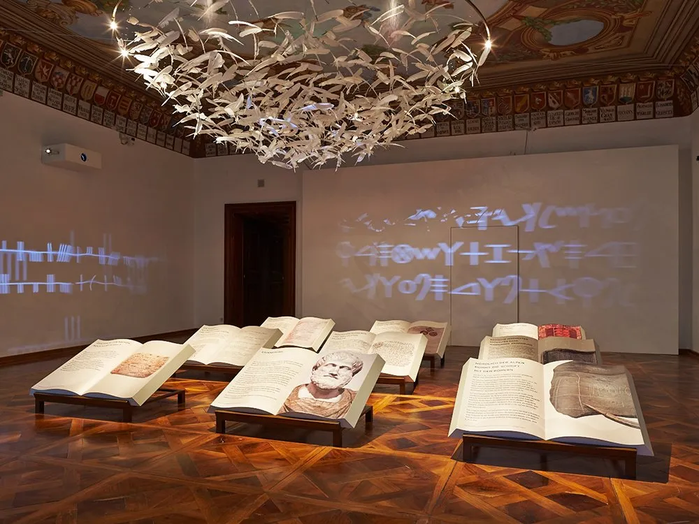 Ausstellungsraum mit offenen Büchern auf Ständern, einer Deckeninstallation mit weißen Vögeln und projizierten Schriftzeichen an der Wand
