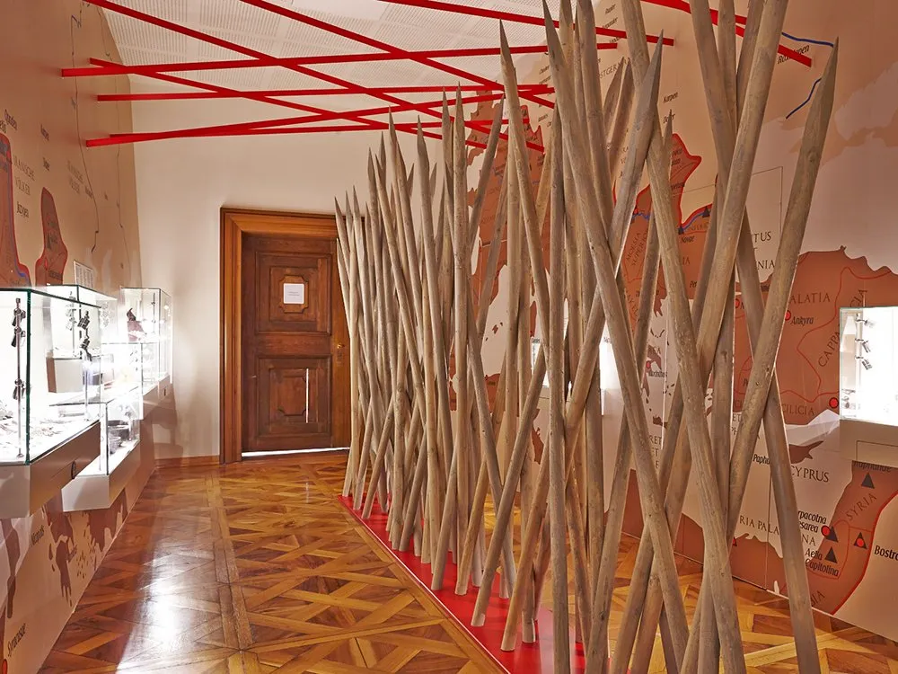 Ausstellungsraum mit Vitrinen und Artefakten sowie einer Installation aus hölzernen Stäben auf einer roten Basis