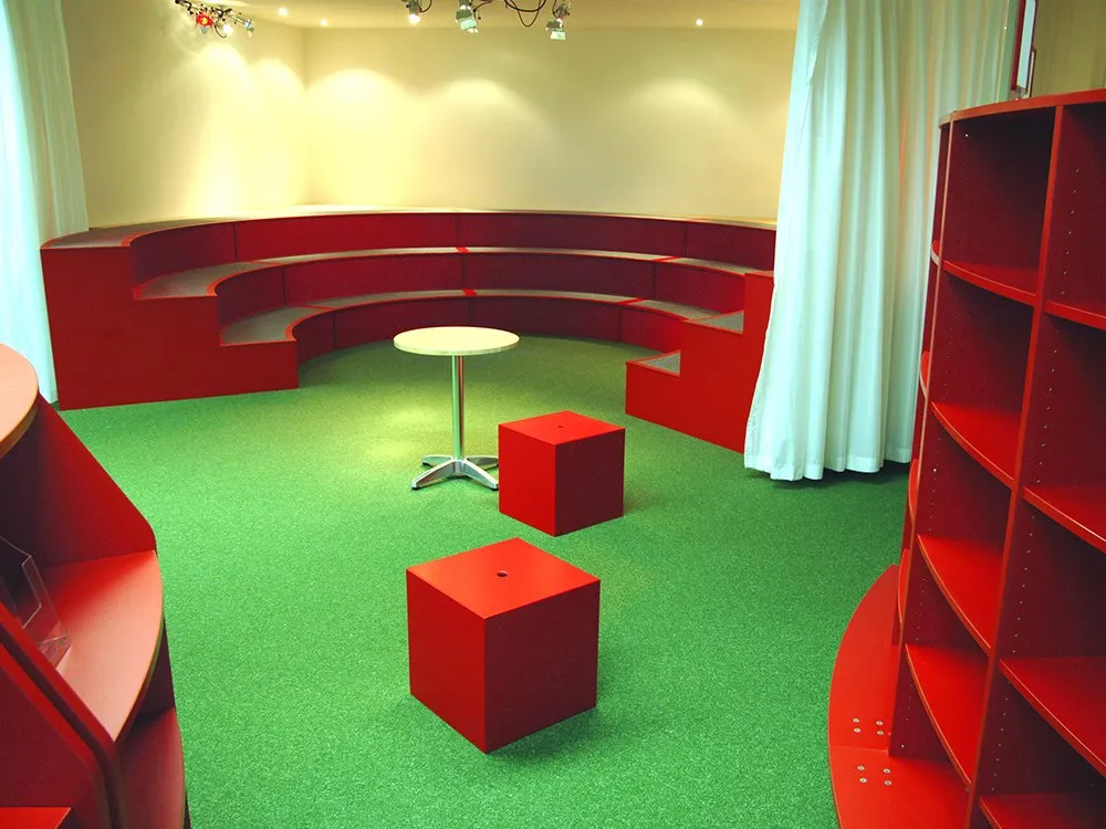 Runde rote Bücherregale mit Sitzfläche, ein runder Holztisch, rote Sitzwürfel und grüner Boden