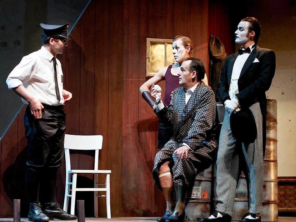 Szene: Polizist, sitzender Mann in einem Kimono und zwei Figuren in auffälliger Kleidung vor einer hölzernen Wandkulisse.