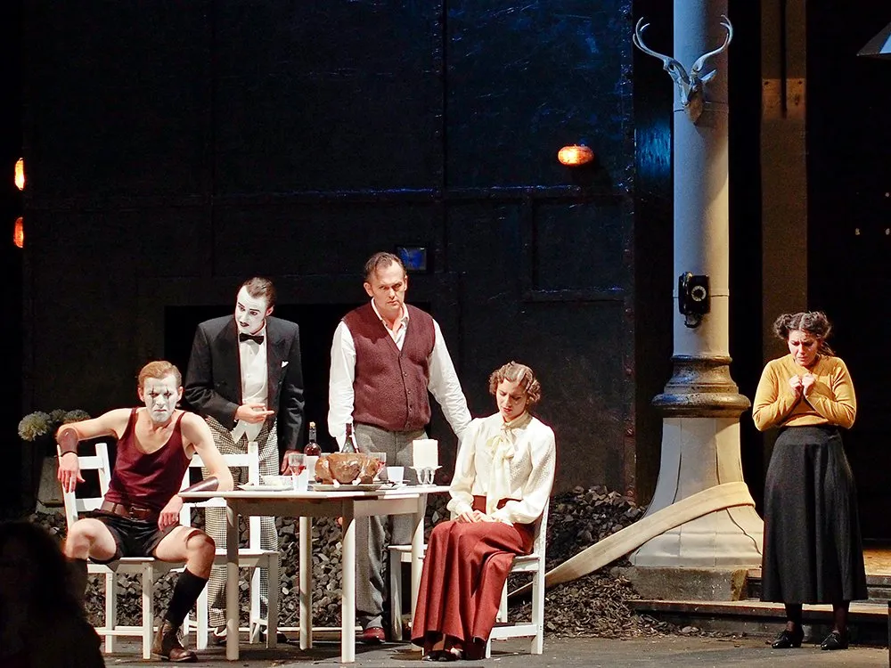 Szene: Zwei Männer mit weißer Gesichtsbemalung, ein Mann in Weste, eine sitzende Frau in rotem Rock und eine stehende Frau in gelber Bluse an einem gedeckten Tisch vor einer dunklen Wandkulisse