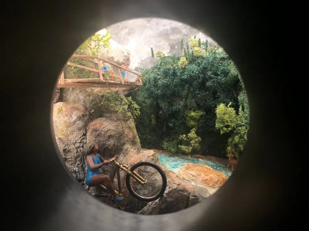 Mini-Diorama mit einer Radfahrerin auf einem Bergpfad, einer Holzbrücke, grünen Pflanzen und einem türkisfarbenen Wasserlauf.